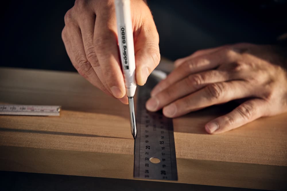 Nahaufnahme der Hände einer Person, die ein Stück Holz mit einem Aluminiumschaft-Maßstab misst und es mit einem edding 8880 Tiefloch Baumarker von edding markiert. Das weiche Licht betont die Präzision und Konzentration bei der Arbeit und deutet auf eine sorgfältige Tischler- oder Handwerkstätigkeit hin.