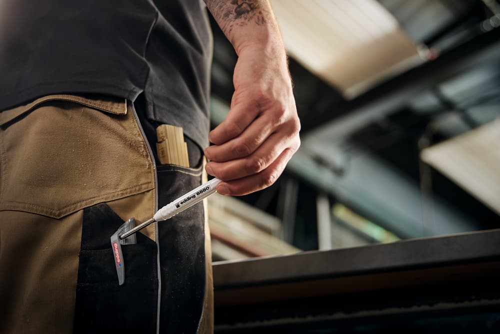 Eine Person in brauner Arbeitshose mit schwarzer Tasche hält einen weißen Druckkugelschreiber mit der Aufschrift „CAT“ in einer Hand. Im Hintergrund ist eine Industriekulisse mit Metallträgern und einer teilweise sichtbaren Werkbank zu sehen, ideal für präzise Markierungen mit dem robusten Schaft des edding 8880 Tiefloch Baumarker-Sets inklusive Graphitminen.