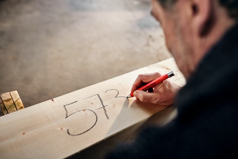 Eine Person schreibt mit einem edding 8890 Automatischer Zimmermannsbleistift HB Zahlen auf ein Holzbrett. Auf dem Brett ist die Zahl „573“ zu sehen. Der Hintergrund ist verschwommen, wodurch der Fokus auf die Hand und das Brett gelegt wird.