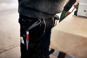 Eine Person in schwarzer Hose ist bei der Arbeit mit Holzbearbeitungsgeräten zu sehen. Werkzeuge, darunter ein roter Edding 8890 Automatischer Zimmermannsbleistift HB und ein kleiner Gegenstand, der einem Klebestift ähnelt, sind in einem an der Hose befestigten Halter verstaut. Sägespäne sind auf der Hose und in der Luft sichtbar.