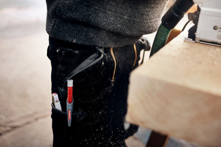 Eine Person in schwarzer Hose ist bei der Arbeit mit Holzbearbeitungsgeräten zu sehen. Werkzeuge, darunter ein roter Edding 8890 Automatischer Zimmermannsbleistift HB und ein kleiner Gegenstand, der einem Klebestift ähnelt, sind in einem an der Hose befestigten Halter verstaut. Sägespäne sind auf der Hose und in der Luft sichtbar.