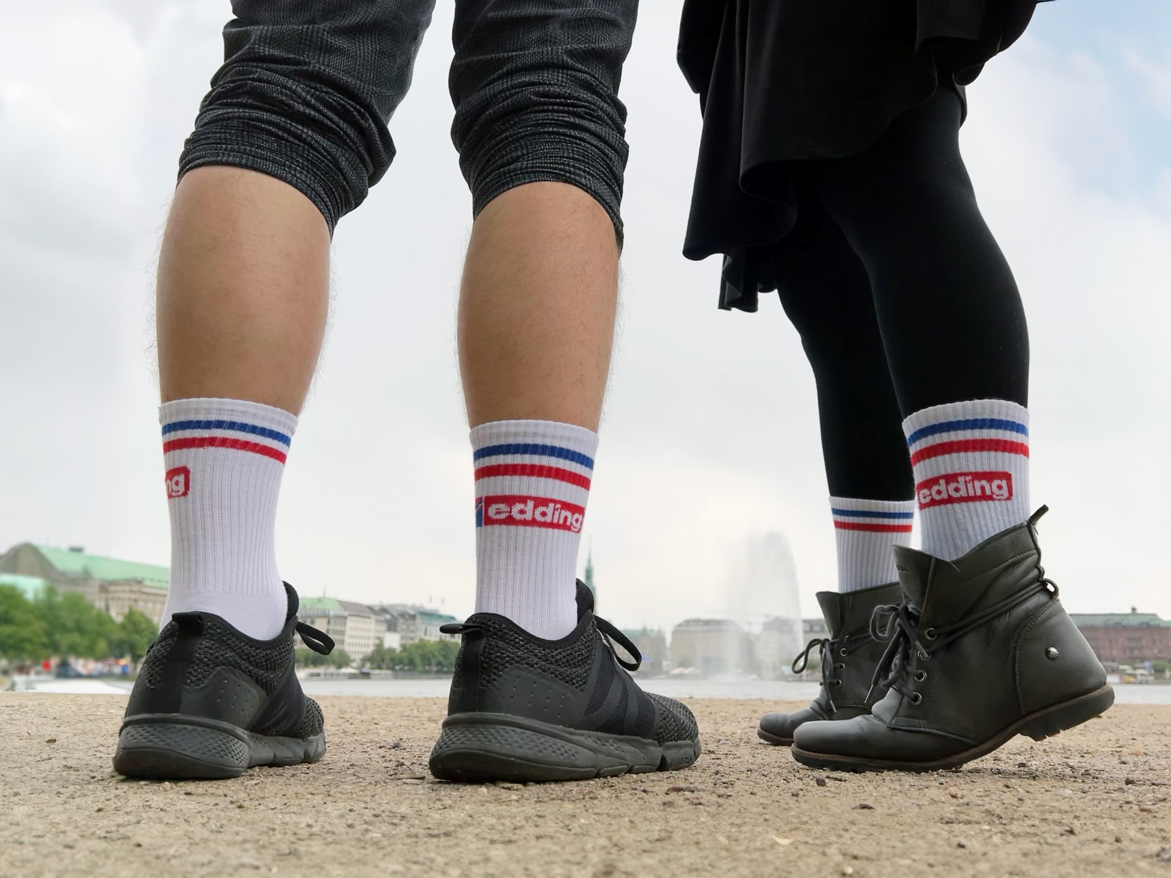 Es sind zwei Paar Beine zu sehen, eines in schwarzen Sportschuhen und kurzen Hosen, das andere in schwarzen Stiefeln und einem Rock. Beide Personen tragen edding Tennissocken mit roten und blauen Streifen am oberen Rand. Diese in Deutschland hergestellten Unisex-Socken verleihen ihnen einen stilvollen Touch, wenn sie draußen in der Nähe eines Gewässers stehen.