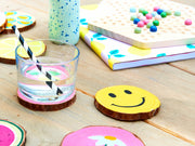 Eine sommerliche Tischdekoration mit einem Glas mit gestreiftem Strohhalm, bunten Untersetzern mit Smileys und einem verspielten Notizbuch, umgeben von fröhlichen Dekorationen und einem edding 5100 Acrylmarker medium von edding.