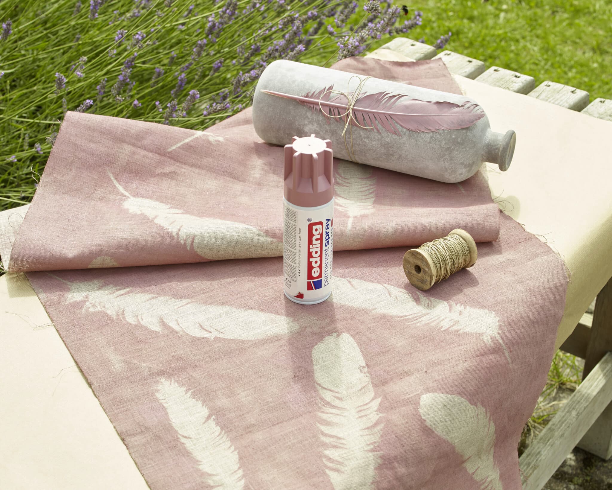 Eine ruhige Bastelumgebung im Freien mit einer Rolle rosa Stoff mit weißem Federmuster neben einer Dose edding 5200 Permanentspray Acryllack Pastell & sanfte Farben, einer Spule Bindfaden und einem dekorativen Kissen.