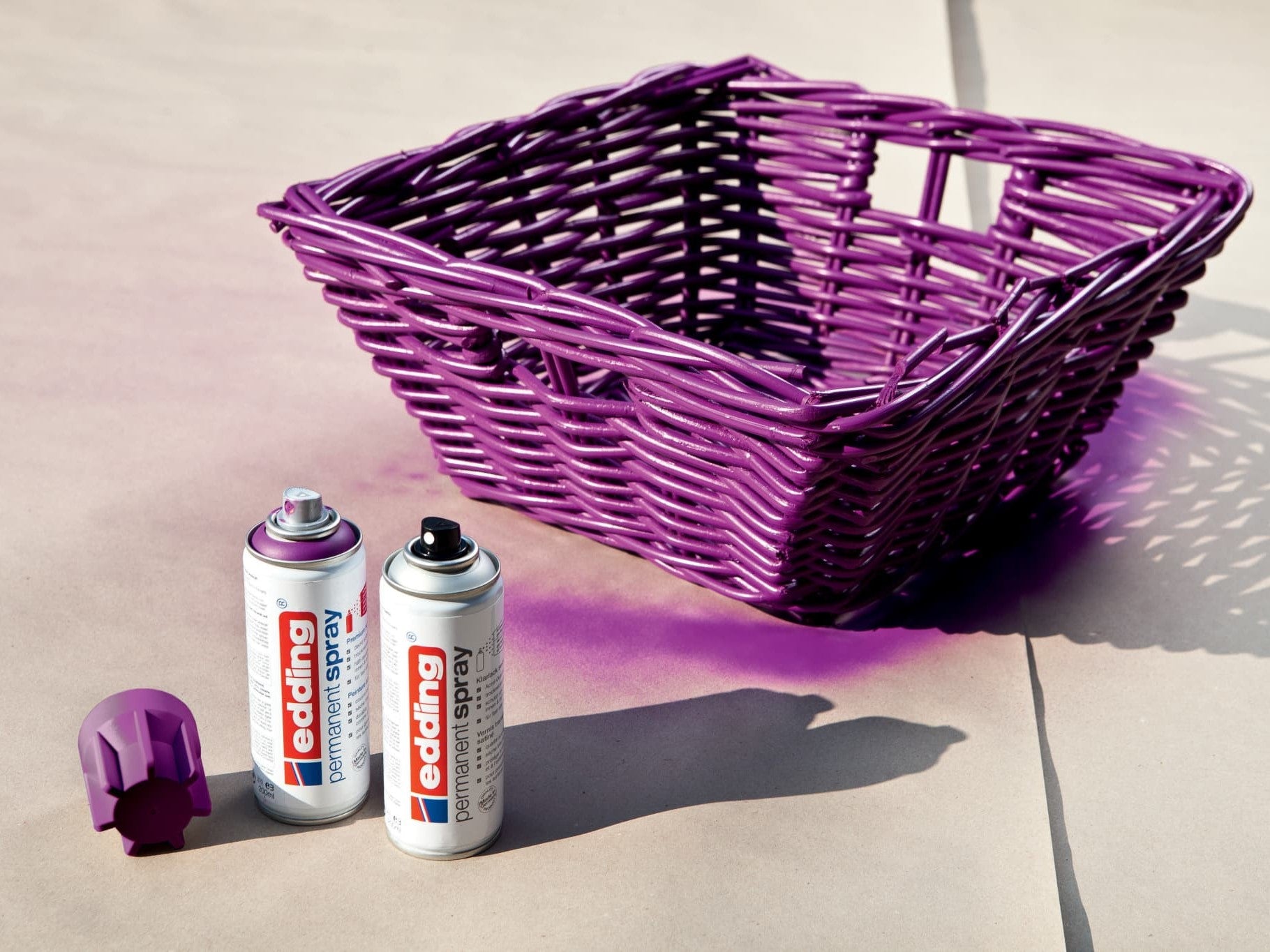Ein violetter Weidenkorb neben zwei abgenommenen Sprühdosen „edding 5200 Permanentspray Acryllack Neon & Kräftige Farben“ und einem kräftigen Neonfarben-Spritzer im Sonnenlicht.