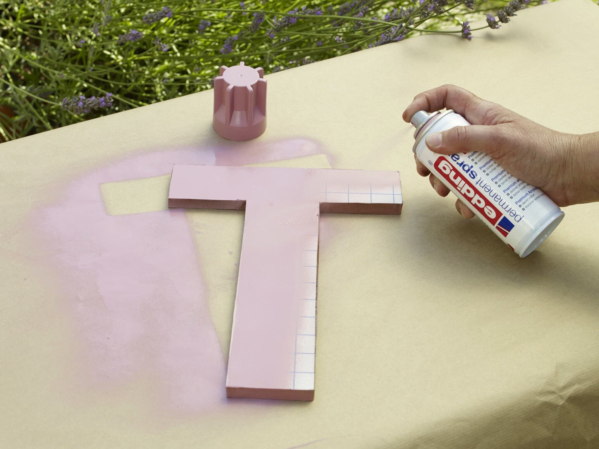 Besprühen Sie einen Holzbuchstaben „t“ mit einem zarten Rosaton und verwenden Sie edding 5200 Permanentspray Acryllack Pastell & sanfte Farben auf einer papierbedeckten Oberfläche im Freien, während eine Hand die Farbsprühdose in Aktion hält.