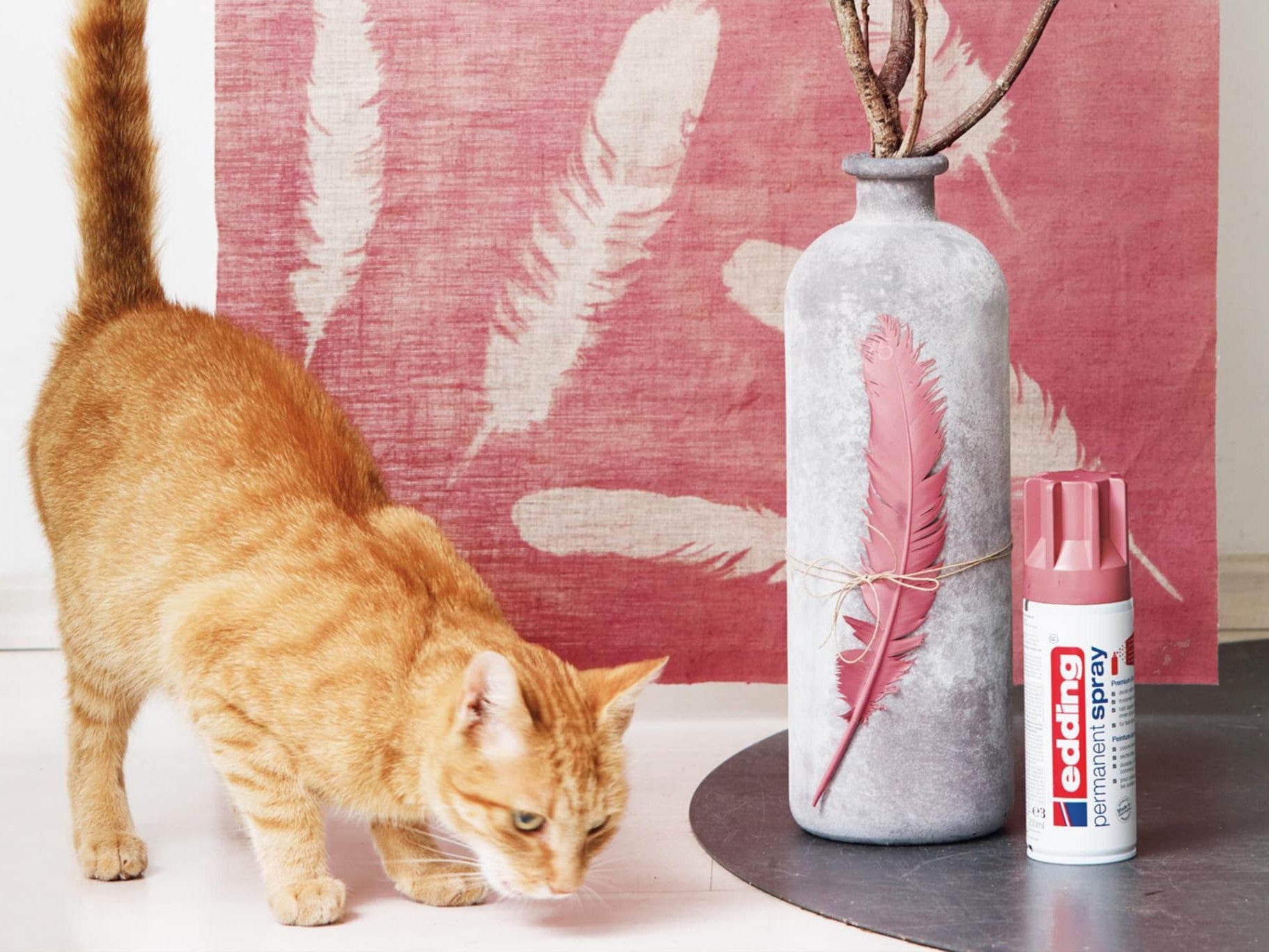 Eine neugierige rote Katze schnuppert an einer grauen Vase, die mit einer roten Feder geschmückt ist. Neben ihr steht eine passende rote Dose „edding 5200 Permanentspray Acryllack Pastell & sanfte Farben“ vor einer großen rosa Leinwand mit Federaufdruck.