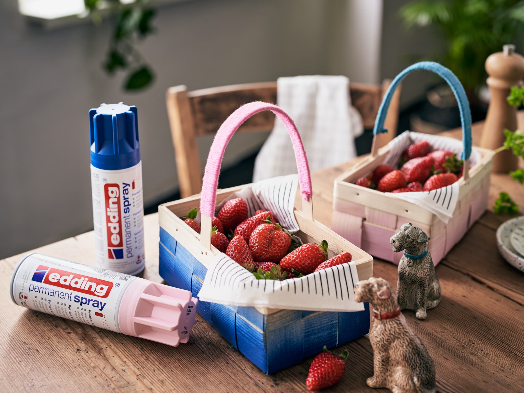 Frische Erdbeeren in Holzkörben auf einem Tisch mit Dosen edding 5200 Permanentspray Acryllack Pastell & sanfte Farben und kleinen dekorativen Katzenfiguren in der Nähe.