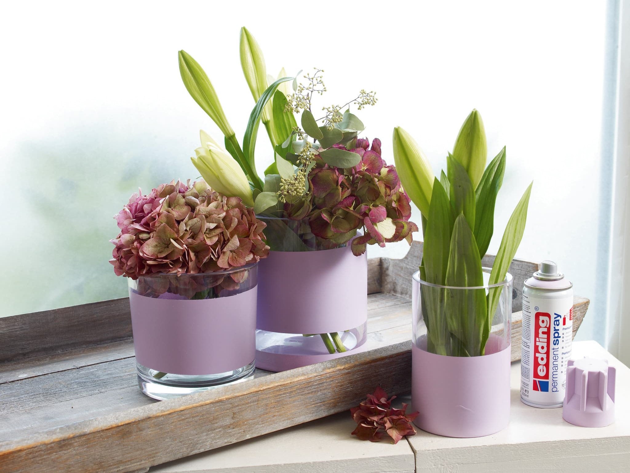 Drei Topfpflanzen mit grünen Blättern und Blüten in verschiedenen Rosa- und Grüntönen, neben einem Fenster auf einer Holzoberfläche platziert, mit einer Dose edding 5200 Permanentspray Acryllack Pastell & sanfte Farben und einem Pinsel.