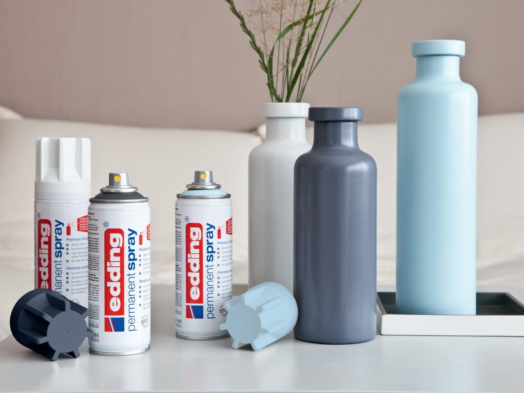 Eine Sammlung von edding 5200 Permanentspray Acryllack Pastell & sanfte Farben-Sprühdosen neben frisch bemalten Flaschen in passenden pastellblauen Farbtönen, präsentiert in einer modernen Inneneinrichtung.