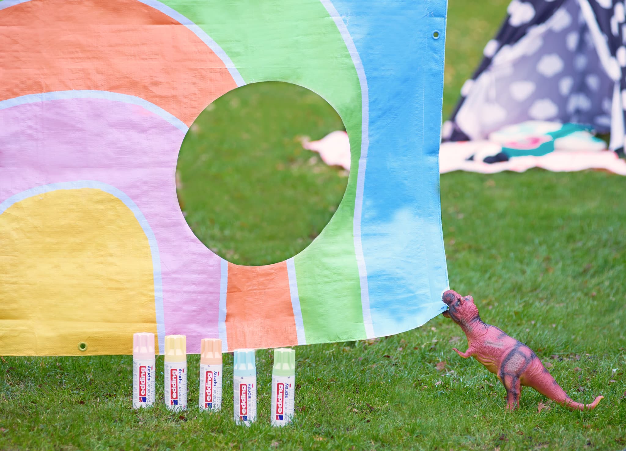 Ein farbenfroher Spieltunnel mit edding 5200 Permanentspray Acryllack Pastell & sanfte Farben auf Gras mit einem süßen Spielzeugdinosaurier, der neben einer Anordnung von Sonnenschutzflaschen steht.