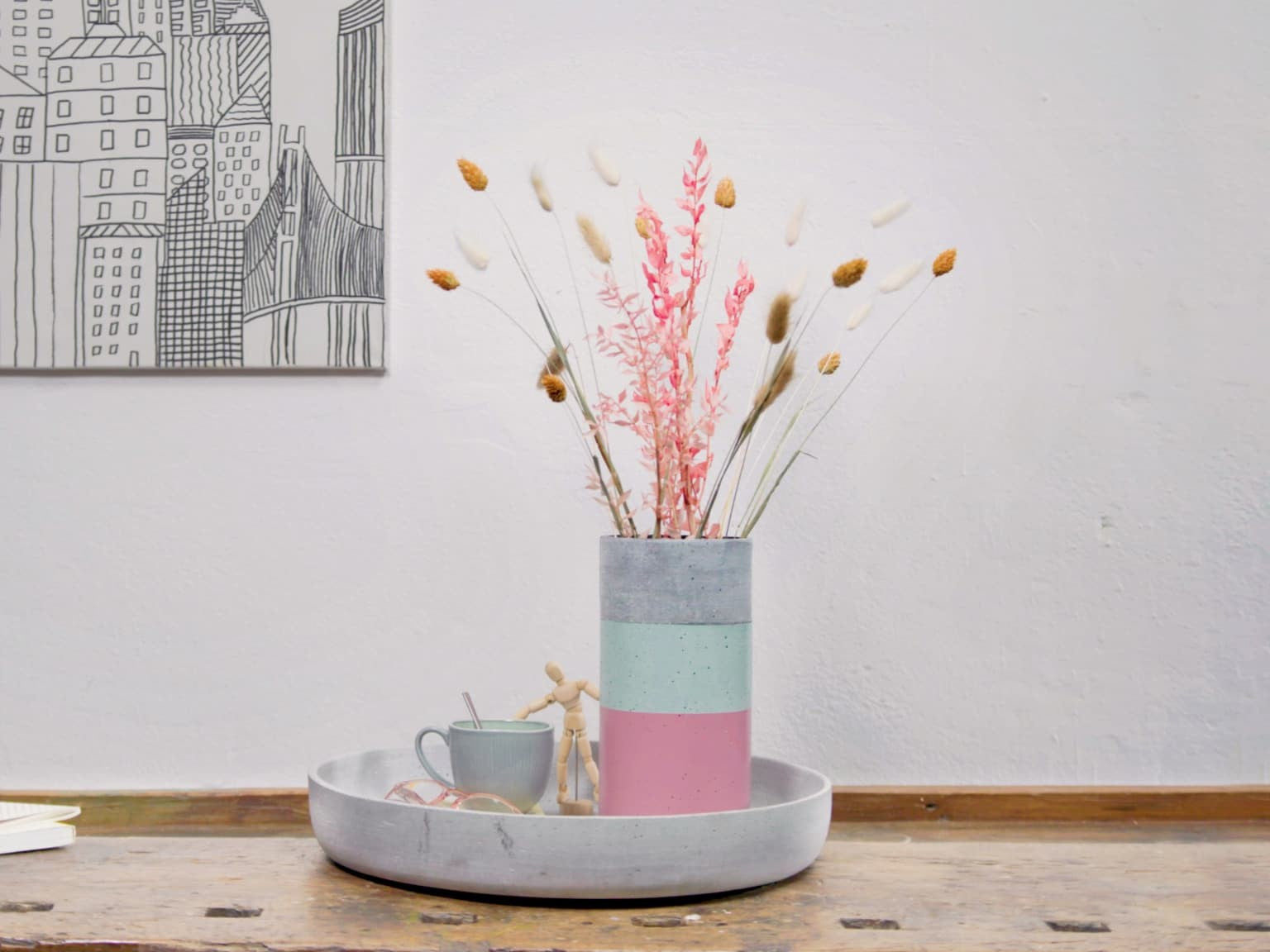 Eine moderne Vase aus edding 5200 Permanentspray Acryllack Pastell & sanfte Farben mit einer Auswahl getrockneter Blumen steht auf einem grauen Tablett neben einer Tasse Kaffee, vor dem Hintergrund einer einfachen Strichzeichnung an der Wand.
