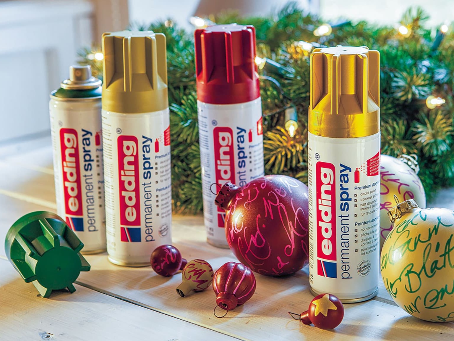 Eine Sammlung von edding 5200 Permanentspray Metallic Acryllack auf einem festlichen Hintergrund mit Weihnachtsdekorationen und Tannenzweigen.