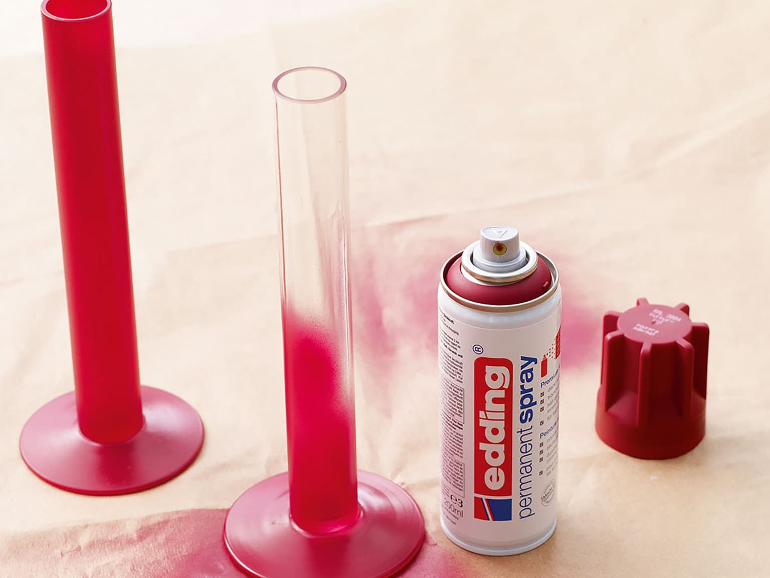 Eine Spraydose edding 5200 Permanentspray Acryllack Neon & Kräftige Farben liegt auf der Seite neben zwei großen roten Kerzenständern auf einer hellen Fläche.