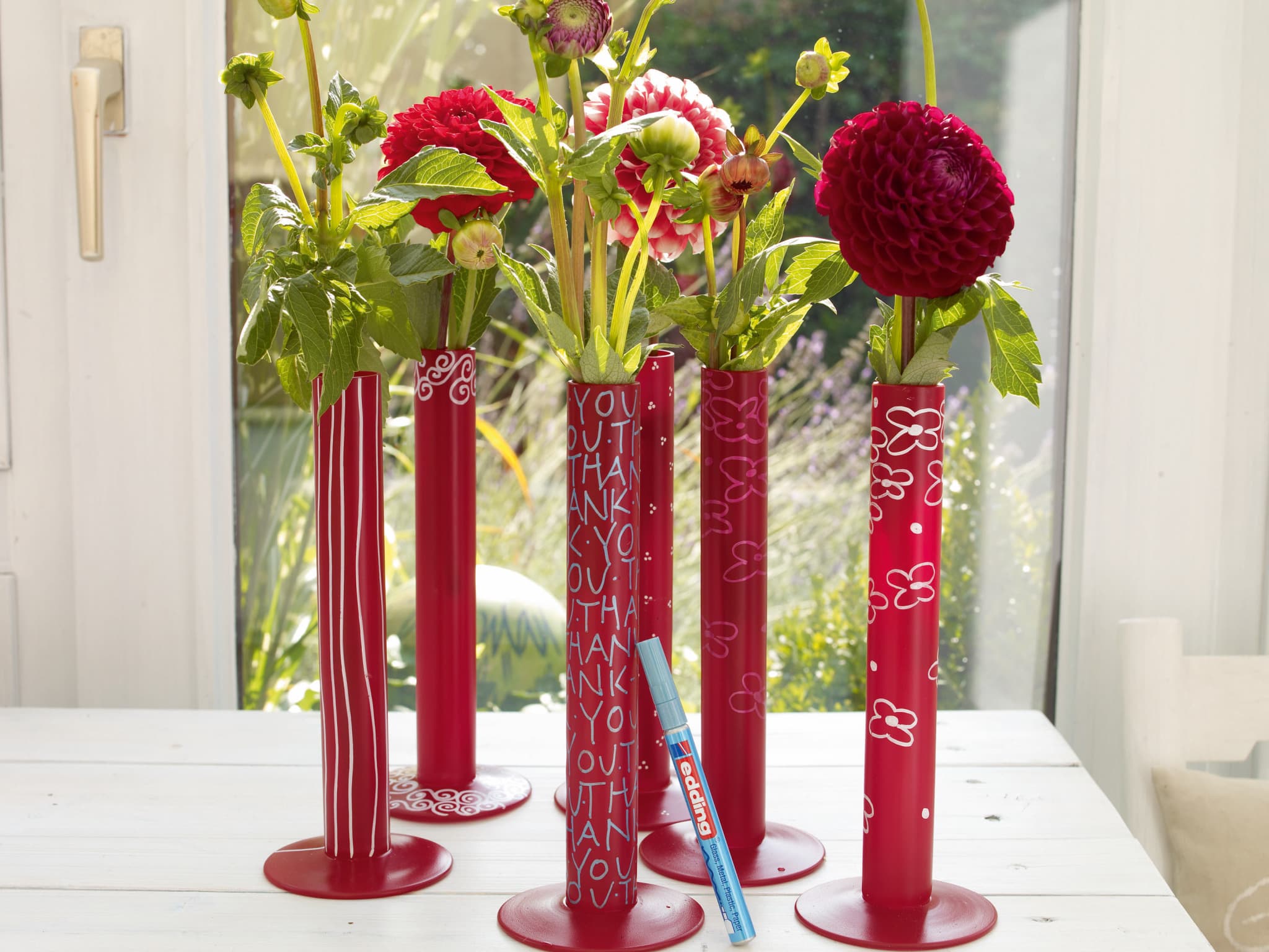 Eine Sammlung leuchtend roter zylindrischer Vasen mit der Aufschrift "Thank You" in Weiß auf der Oberfläche, besprüht mit edding 5200 Permanentspray Acryllack Neon & kräftige Farben für ein glänzendes Finish, gefüllt mit einer Auswahl an wunderschönen roten
