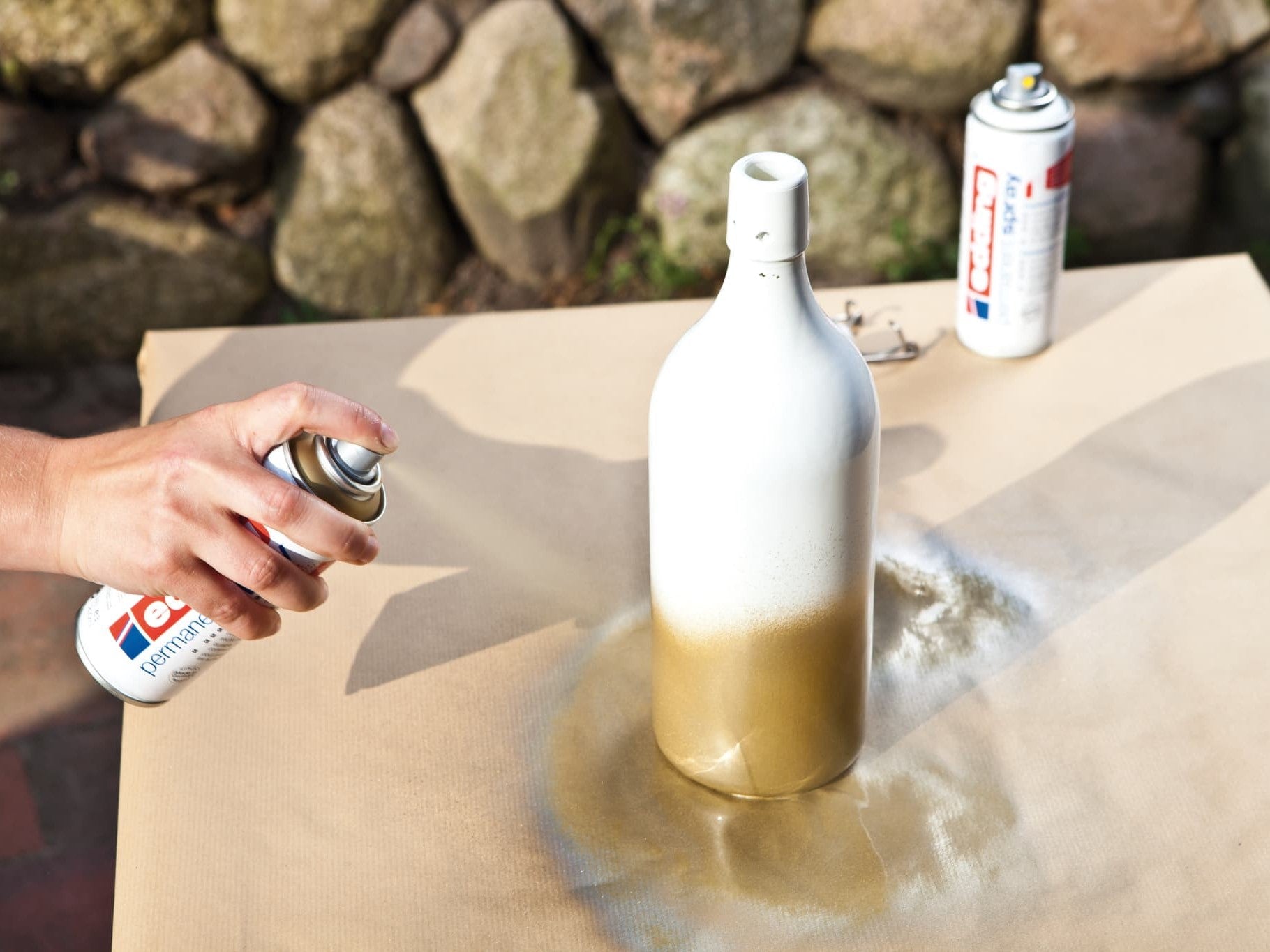 Eine Flasche mit edding 5200 Permanentspray Metallic Acryllack auf einer papierbespannten Fläche im Außenbereich gold besprühen.
