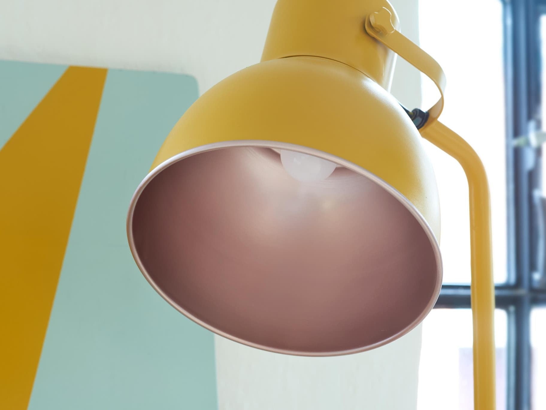 Eine moderne gelbe Schreibtischlampe mit rosafarbenem Innenschirm ist an einer Wand mit geometrischen Mustern in blassen Tönen montiert und weist eine mit edding 5200 Permanentspray Metallic Acryllack veredelte Oberfläche auf.