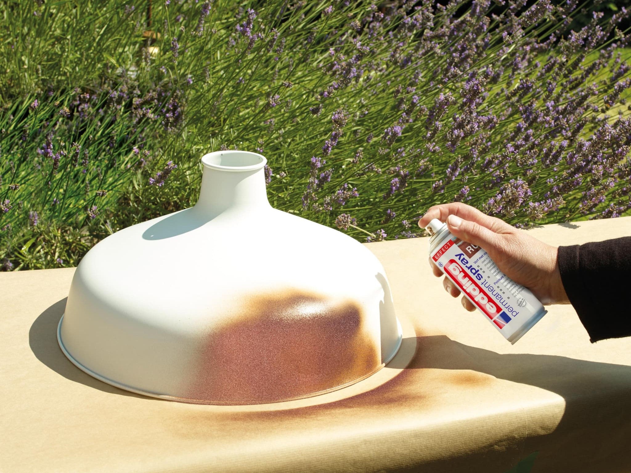 Eine Hand trägt eine Schicht edding 5200 Permanentspray Effekt Acryllack auf eine Vase auf, die an einem sonnigen Tag im Freien vor einem Hintergrund aus Grün- und Lavendelpflanzen steht.