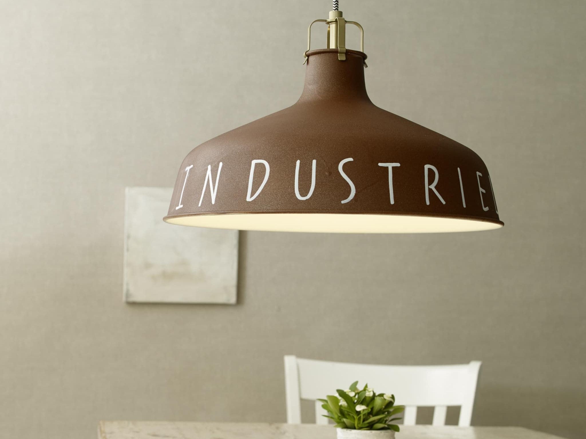 Eine Pendelleuchte im Industriestil von edding mit dem Wort „Industrie“ darauf, die über einem schlichten Tisch mit einer kleinen grünen Pflanze an einer neutralen Wand hängt. Der unverwechselbare Look der Lampe