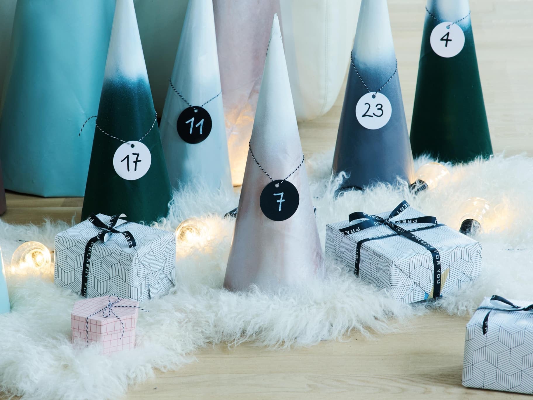 Ein Adventskalender mit nummerierten kegelförmigen Bäumen und elegant verpackten Geschenkboxen, die auf einer mit Edding akzentuierten weißen Oberfläche ruhen und mit sanftem Licht beleuchtet werden.