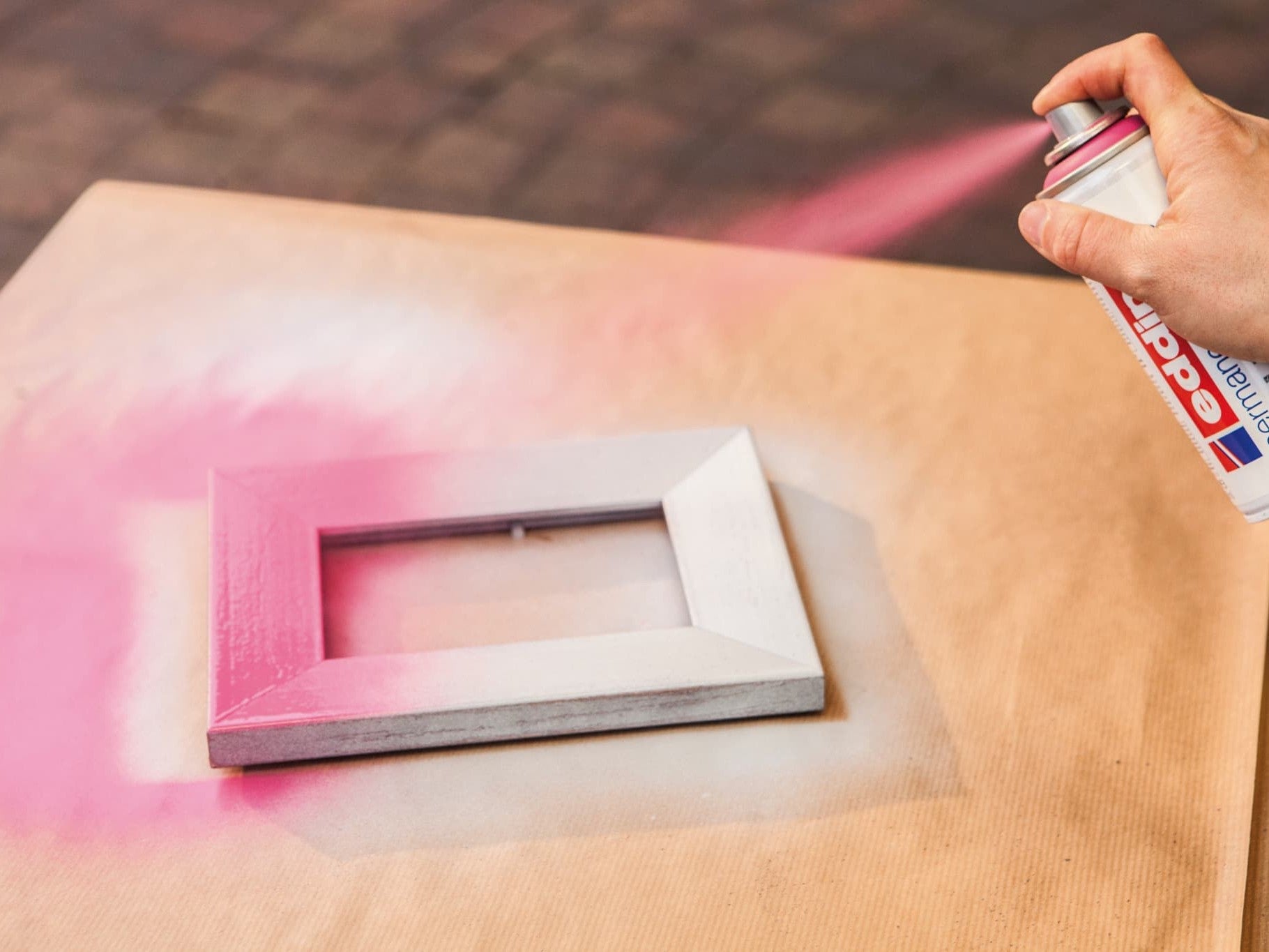 Eine Hand trägt den Klebstoff edding 5200 Permanentspray Acryllack Neon & Kräftige Farben auf einen kleinen Holzrahmen vor einem rosa Overspray-Hintergrund auf einem Arbeitstisch auf.