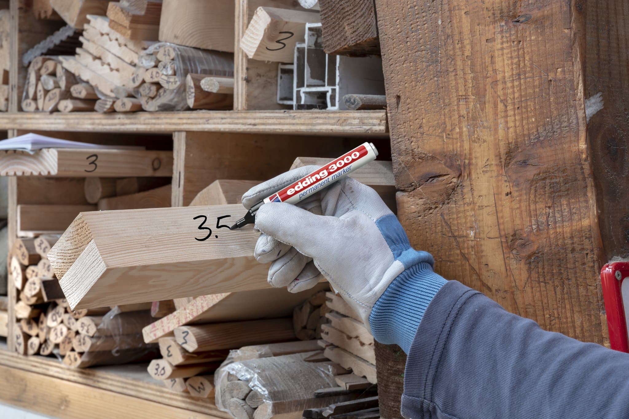 Eine Person in einem Handschuh markiert einen Holzbalken mit einem edding 3000 Permanentmarker 8er Dose in einer Holzwerkstatt, im Hintergrund Regale mit beschrifteten Holzstücken.
