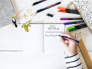 Eine Person beim Handlettering in einem leeren Notizbuch, umgeben von bunten Markern, einem edding 1800 Präzisionsfeinschreiber und niedlichen Schreibwaren auf einer weißen Holzoberfläche.