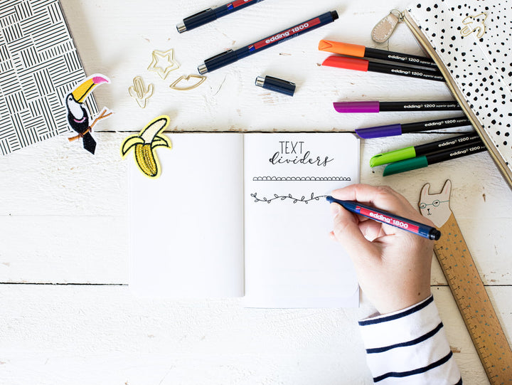 Eine Person beim Handlettering in einem leeren Notizbuch, umgeben von bunten Markern, einem edding 1800 Präzisionsfeinschreiber und niedlichen Schreibwaren auf einer weißen Holzoberfläche.