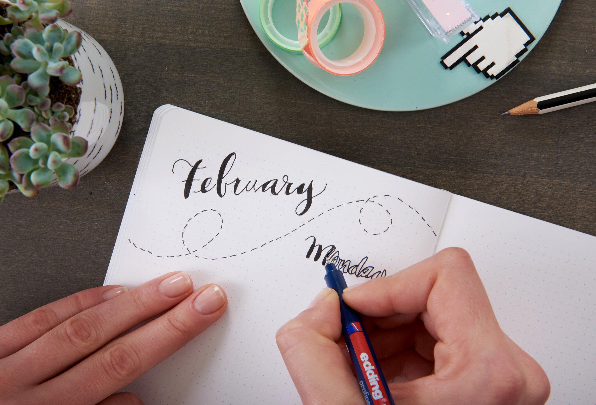 Eine Person übt kreatives Handlettering in einem Notizbuch, wobei das Wort „Februar“ in eleganter Schrift mit einem edding Fineliner geschrieben ist.
