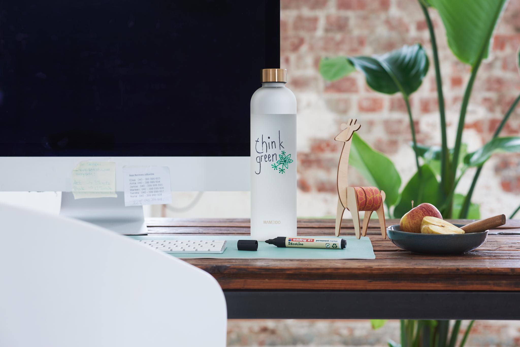 Ein moderner Arbeitsbereich mit einem eleganten Computermonitor auf einem Holzschreibtisch, ausgestattet mit einer edding „Think Green“-Wasserflasche, einer dekorativen Giraffenfigur aus Holz und einer Schale mit frischem Obst daneben.