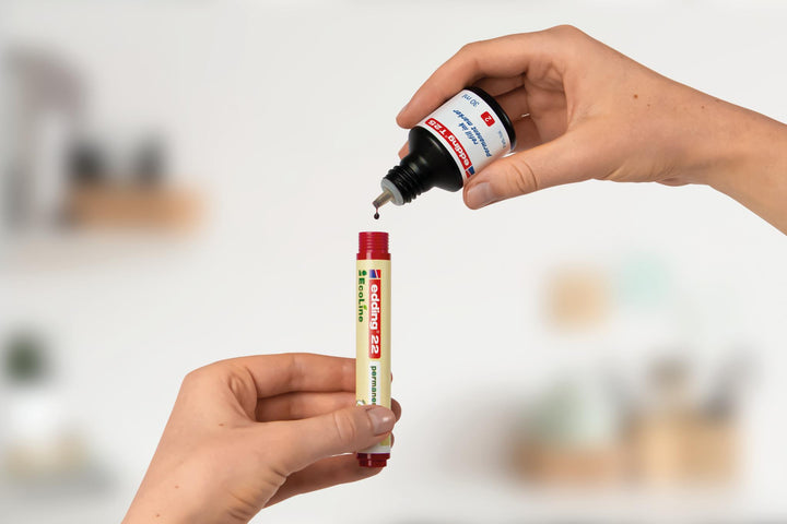 Auftragen von Klebstoff aus einer Quetschflasche auf die Spitze eines edding 21 EcoLine Permanentmarker 4er-Sets vor einem unscharfen Raumhintergrund.