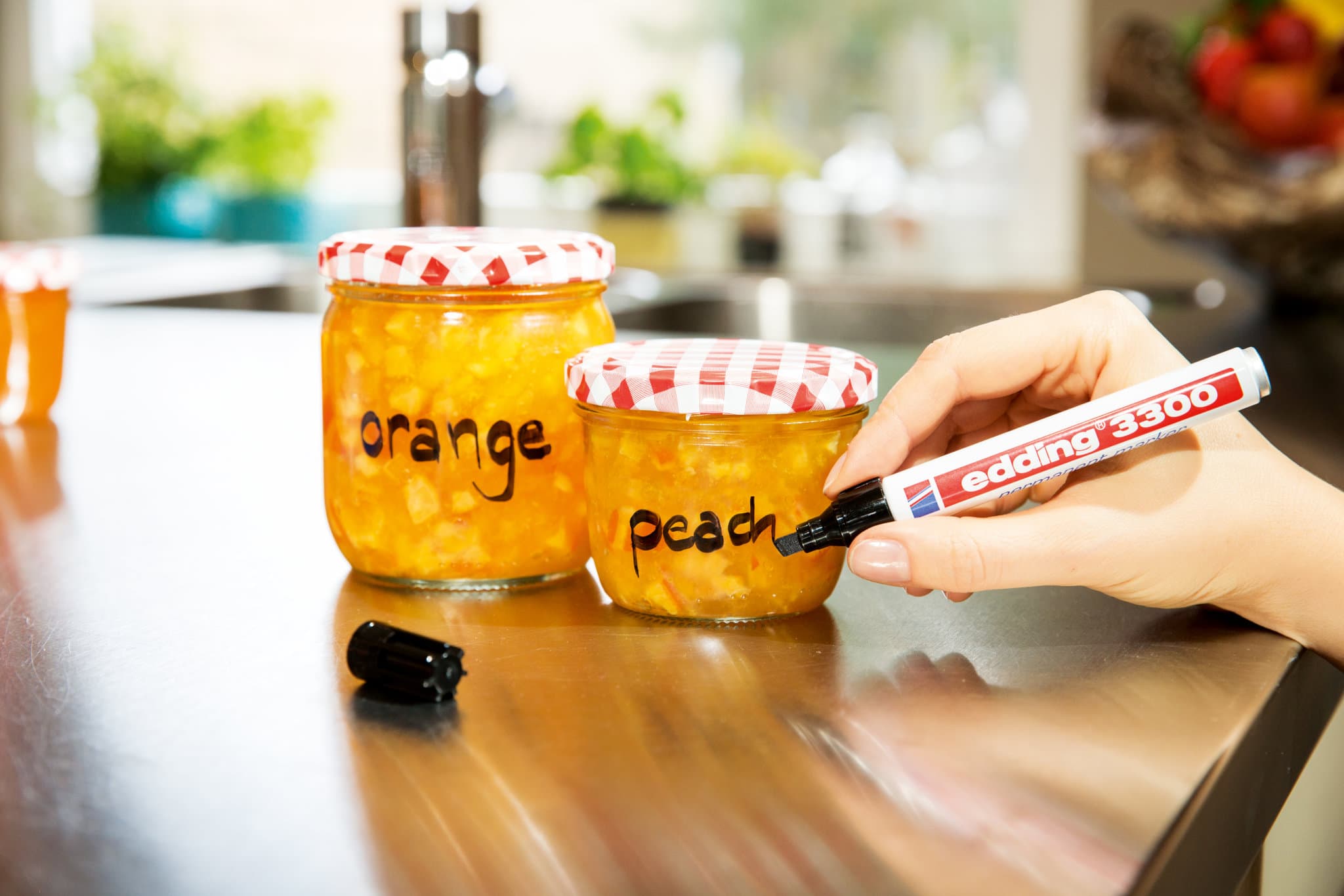 Eine Person beschriftet selbstgemachte Gläser mit Orangen- und Pfirsichmarmelade mit einem Permanentmarker und verwendet dabei den edding 3300 Permanentmarker für farbintensive Tinte.