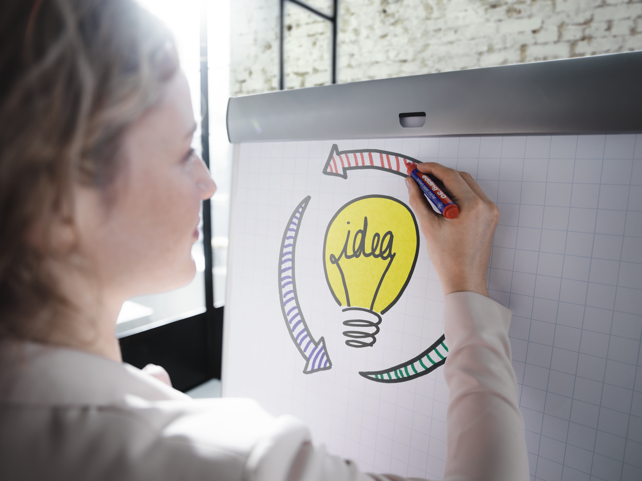 Eine berufstätige Frau zeichnet mit einem edding 33 Brillant-Papiermarker eine farbenfrohe Ideenglühbirne auf ein Whiteboard, die Kreativität und Brainstorming symbolisiert.
