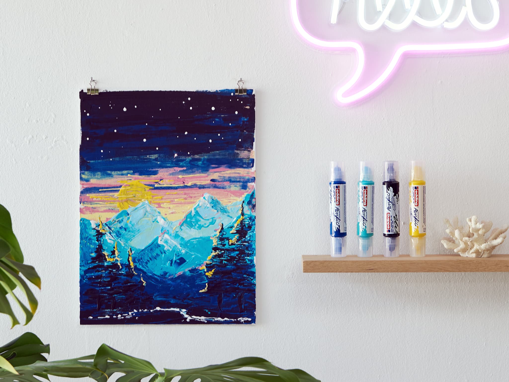Eine lebhaft gemalte Bergszene, erstellt mit dem 5400 Acryl 3D Double Liner 5er-Set von edding, hängt an einer weißen Wand und setzt einen Farbtupfer neben einem Neonlichtschild mit einer Reihe von
